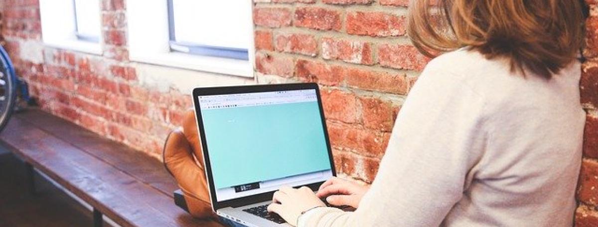 A woman sits along a bench and types on her laptop.