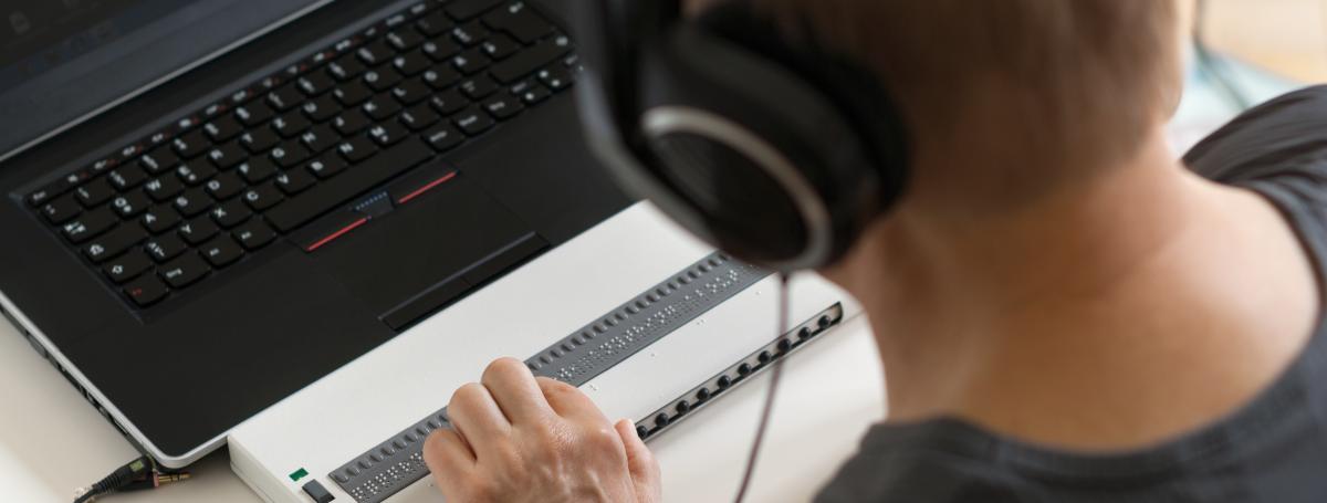 A man is working on laptop computer with a refreshable braille display. He is wearing headphones.