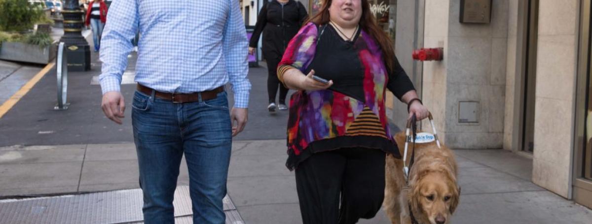 A woman walks down a busy city sidewalk with a young man. She is walking alongside her guide dog in harness.