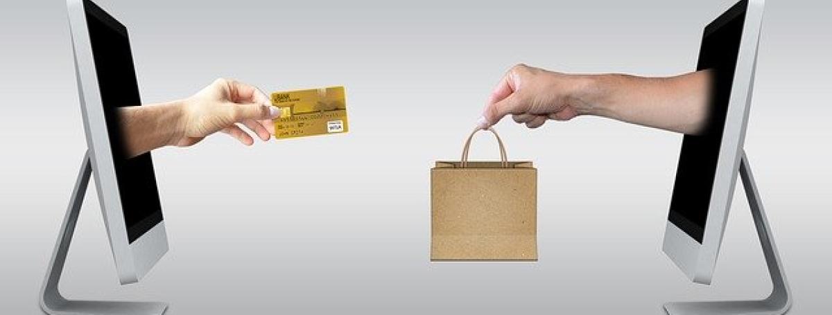 A photoshopped image of two computer monitors facing each other. From the screen of monitor on the left, a hand extends holding a credit card. From the screen of monitor on the right, a hand extends holding a shopping bag.
