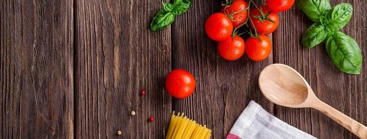 A cutting board. A wooden spoon, basil, tomatoes and pasta sit on top of the board.