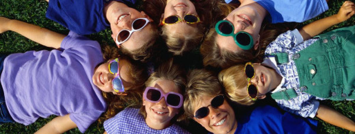 A circle of children laying on the grass looking up. They are all smiling with colourful sunglasses.