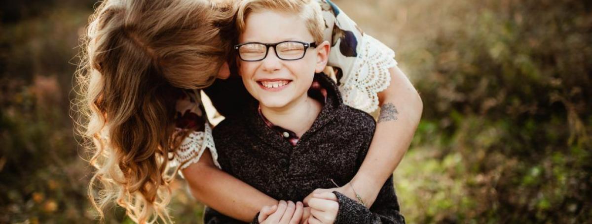 Woman hugging boy wearing glasses outdoors.