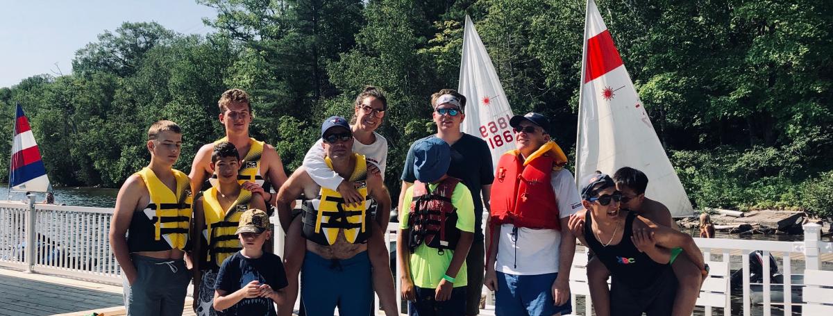 A group of people standing on the dock in shorts and life jackets. They are smiling and in some cases piggy-backing others.