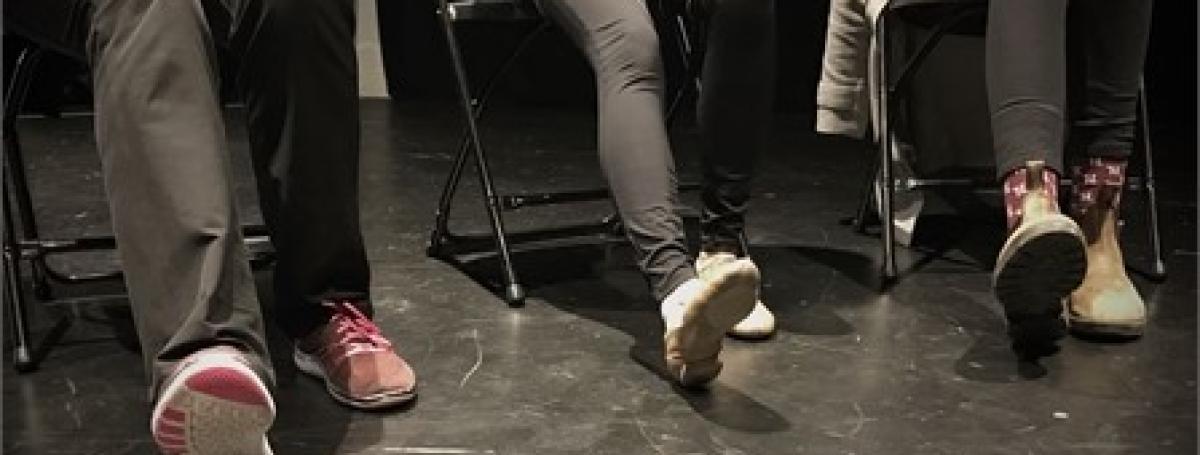 Three people in a dance studio, facing forward, pictured from their hips down. They are sitting on chairs with their right legs extended and their right foot flexed upwards while their heels are on the floor.