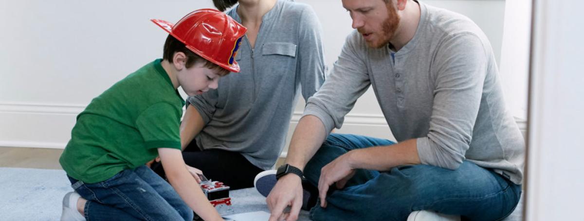 A family sits on the floor looking at a floor plan. There is a red fire extinguisher on the floor and the boy is wearing a firefighter hat.