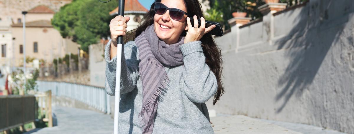 A young woman sits alone outdoors. In her left hand, she is holding her smartphone up to her ear. In her right hand, she holds her white cane