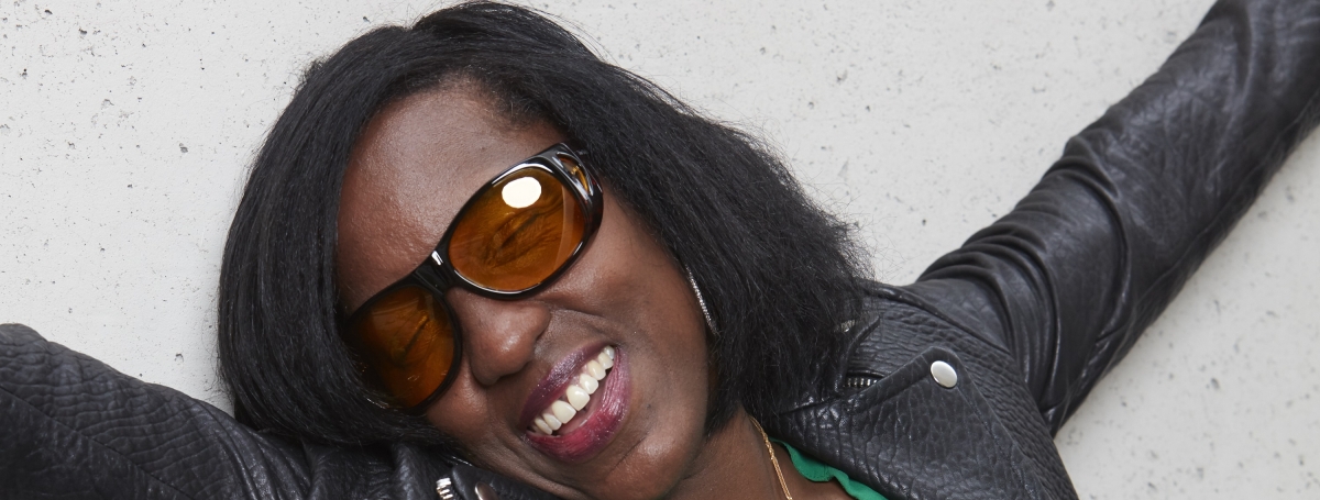 Une femme noire, souriante, portant de grosses lunettes orangées, les bras dans les airs.