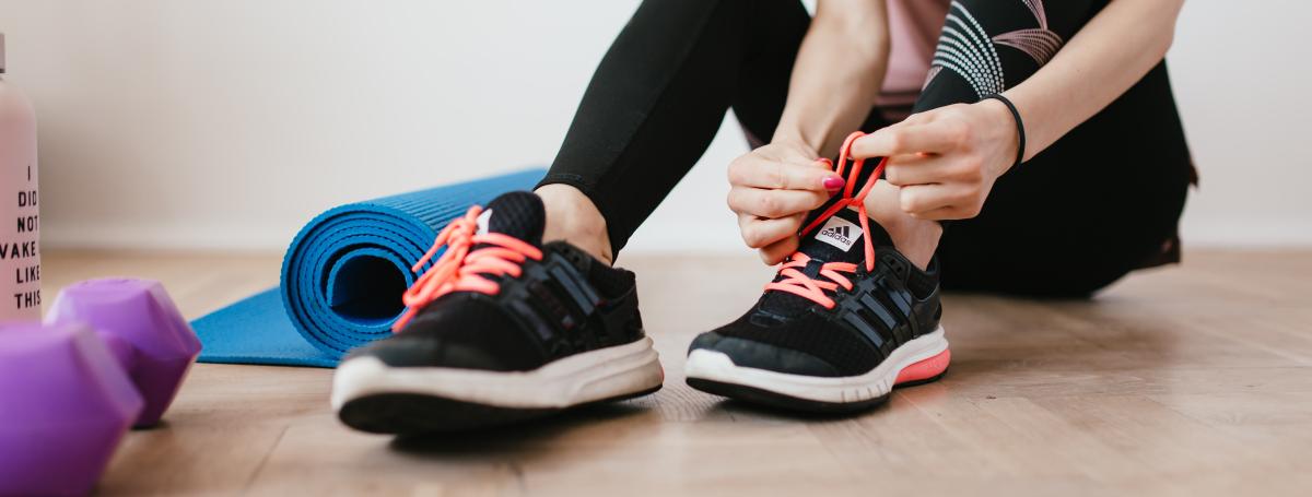 A woman is sitting on the floor tying her running shoes with a yoga mat and dumbbell beside her.
