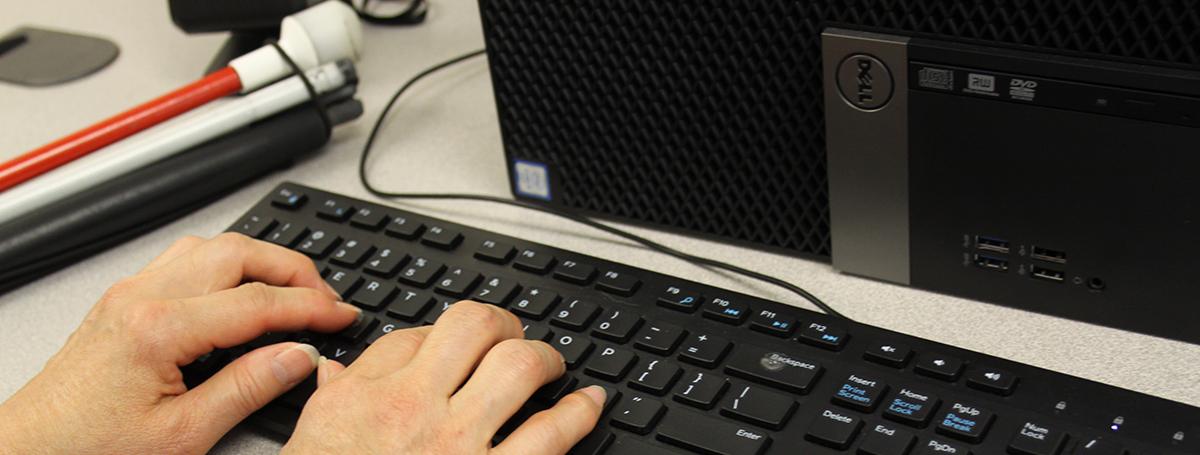 Hands typing on a desktop keyboard. A white cane sits on top of the table to the left of the keyboard.