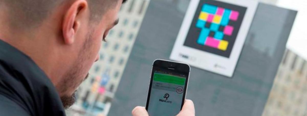 A man holds an iPhone. The NaviLens app. is displayed. He is scanning a QR Code.