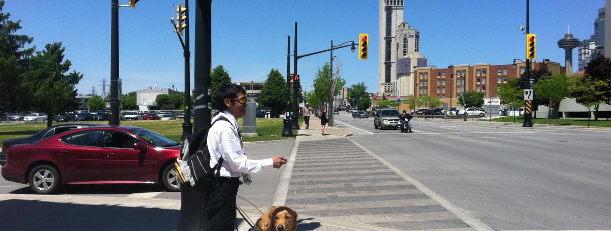 A young man and his guide dog stand on the corner of a busy intersection. In his right hand, he holds a Key2Access Fob. In his left hand, he holds the Guide Dog harness.