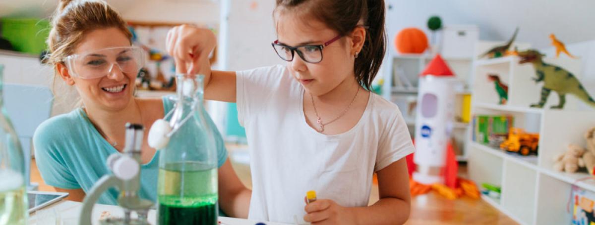 A young girl with glasses stands in front of a table with a microscope and a bottle filled with green liquid.