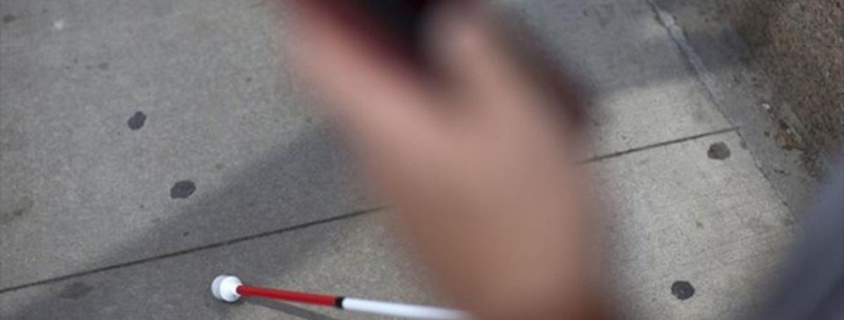 A white cane navigates a sidewalk. A hand holding a smartphone in the foreground.