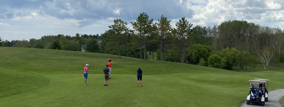 A lush green golf course. Four players stand on the course. A golf cart sits nearby.