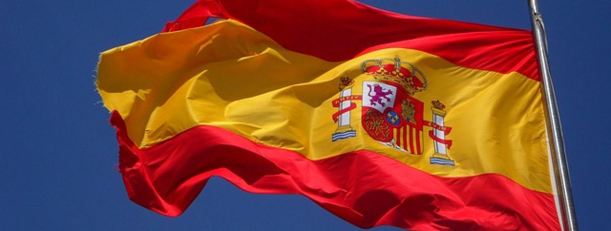 The flag of Spain on a flag post waving in the cloudy blue sky.