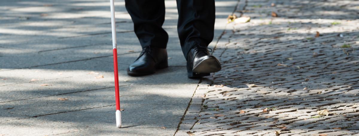 From the knee down, a man's legs walking along a city sidewalk with his white cane.