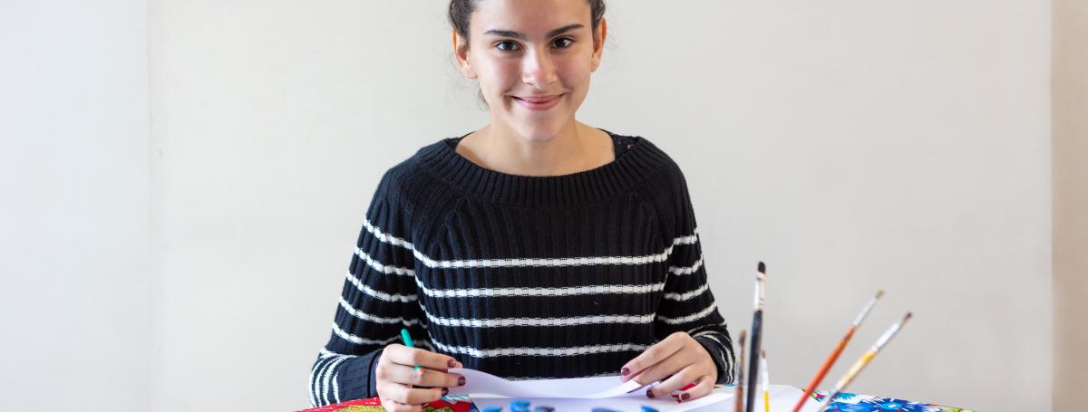 A young woman sits at a table making art with pencils, paper, scissors, magnifying glass, brush and paints in colourful mugs.