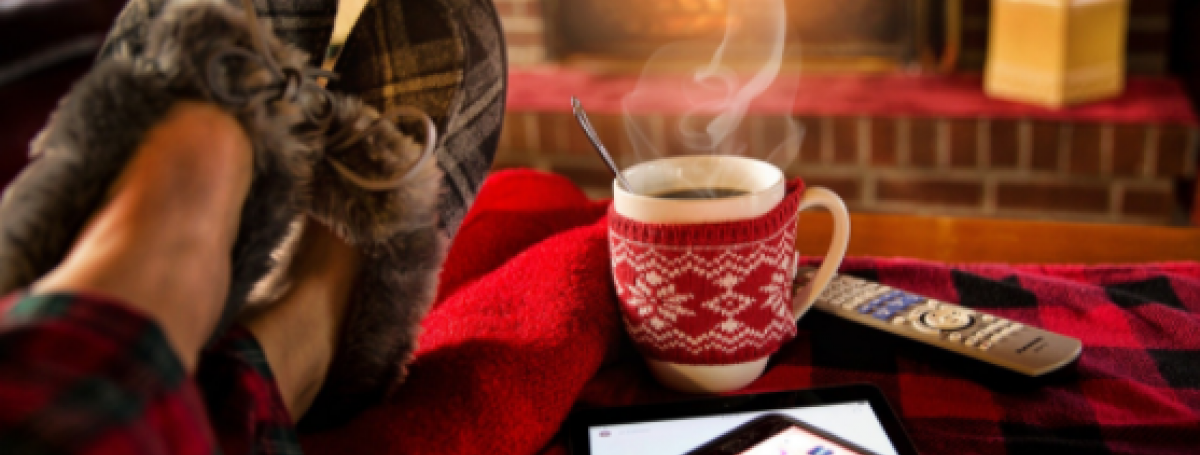 Pieds d'une personne confortablement assise devant un feu de foyer, avec une tasse chaude, des pantoufles et un téléphone intelligent.