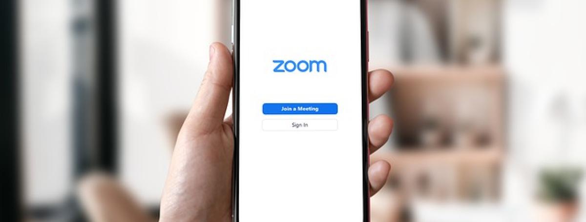A hand holds up an iPhone 11. On the screen, the Zoom app is displayed.