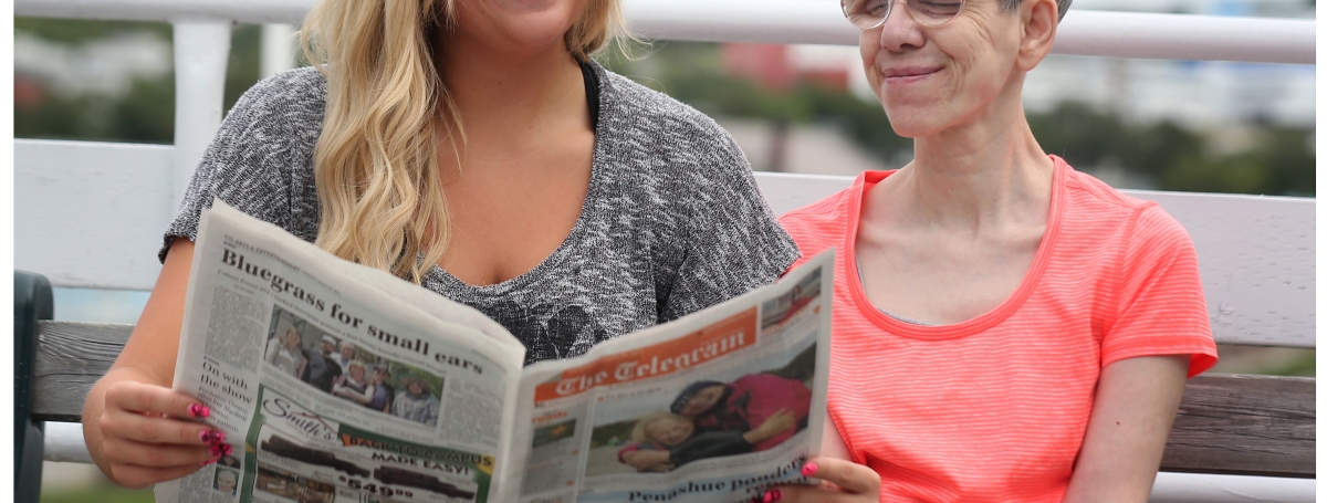 a young woman reads a newspaper to an older woman.