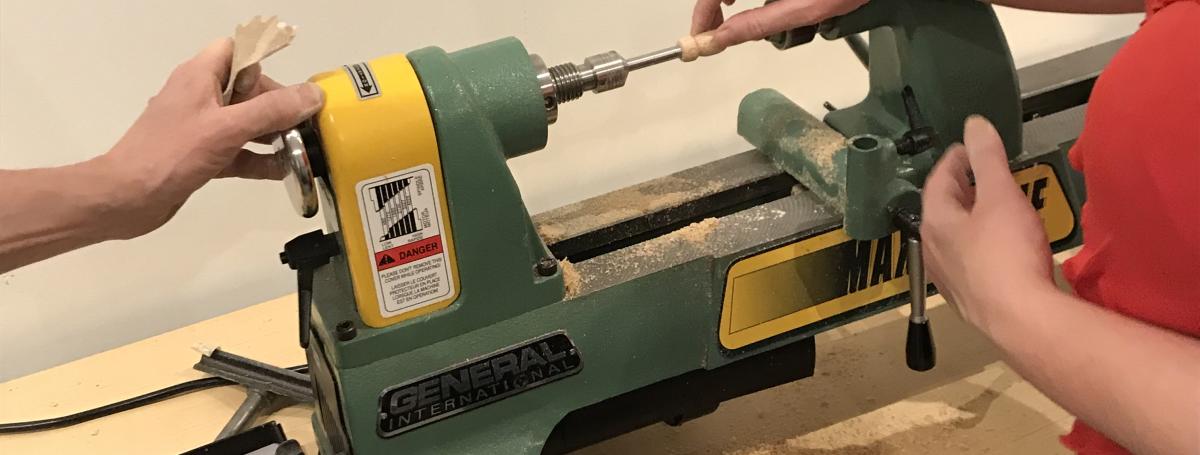 A lathe sits on a counter. Two hands maneuver a piece of wood.  