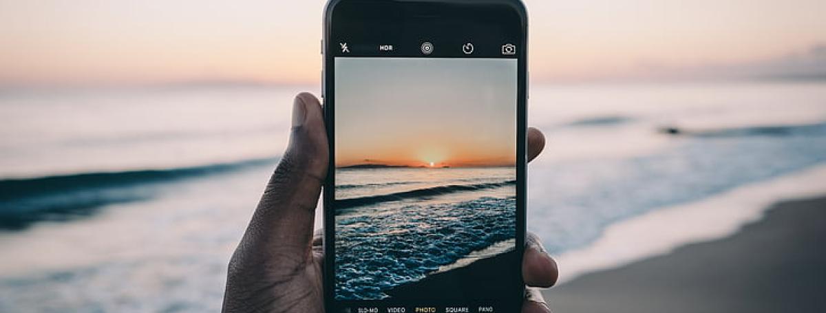 Une main tenant un iPhone devant la mer pour prendre une photo. On voit le paysage sur et derrière l'écran.