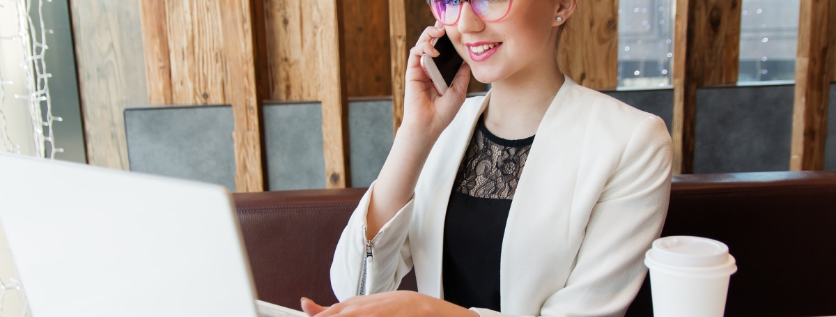 Une jeune femme portant des lunettes parle au téléphone et est sur l'ordi. un café pour emporté est déposé sur la table.