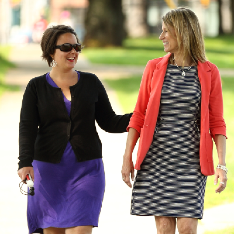 An example of the sighted guide technique. Two woman walk down a sidewalk together. A woman holds the guide's arm lightly above the elbow and allows the guide to walk one-half step ahead.