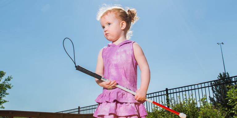 Little girl stands on tactile marker holding her white cane. She's wearing a purple dress and matching shoes.