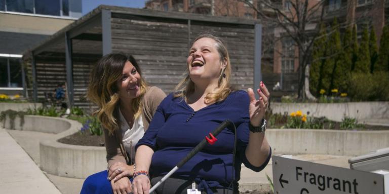 Une femme brune assise souriante et attentive aux côtés d'une femme blonde souriante tenant une canne blanche souriante. Elles sont toutes deux assises à côté d'un jardin olfactif. 