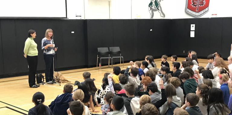 An ambassador stands at the front of a room and gives a presentation to 30+ seated children.