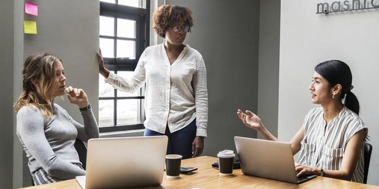 Three women in a meeting. Two women are sitting at a table in front of a laptop. Another woman stands near a window. They are engaging in a serious conversation.
