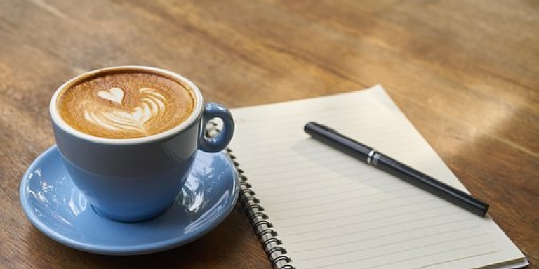 Une tasse de café avec un cœur dessiné dans le lait, sur un table en bois, à côté d'un cahier de note et d'un crayon.