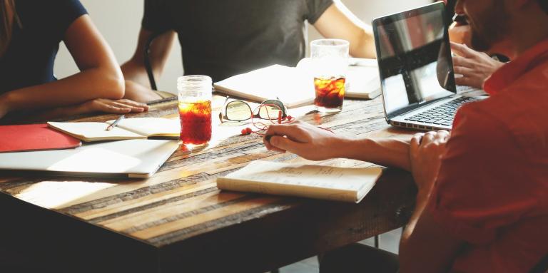 an extreme close up of 4 people sitting at a wooden table. A laptop, paper, pens, glasses, notebooks are infront of them