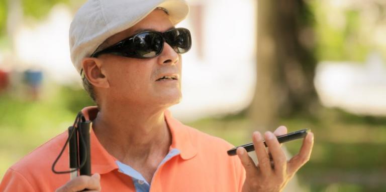 A man wearing sunglasses and a hat holds an iPhone in his left hand. A white cane sits in his right hand.