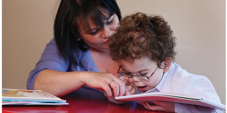 A young boy reads a tactile book. His mother helps guide his hands over the book.