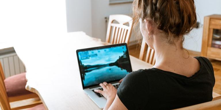 The back of a young woman's head. She sits at kitchen table and types on her laptop computer.