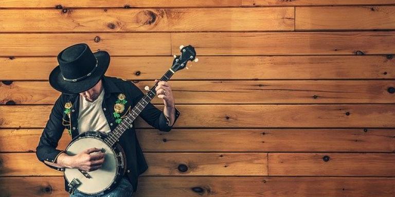 A male musician leans against a wall while strumming on a banjo.