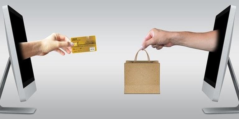 A photoshopped image of two computer monitors facing each other. From the screen of monitor on the left, a hand extends holding a credit card. From the screen of monitor on the right, a hand extends holding a shopping bag.
