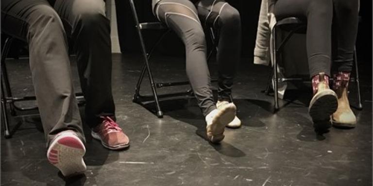 Three people in a dance studio, facing forward, pictured from their hips down. They are sitting on chairs with their right legs extended and their right foot flexed upwards while their heels are on the floor.