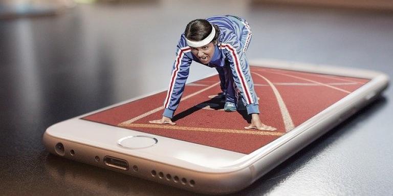 An iPhone sits on a table top. Superimposed on top of the phone is a funny photoshopped picture of a miniature/shrunken woman about to run a marathon.