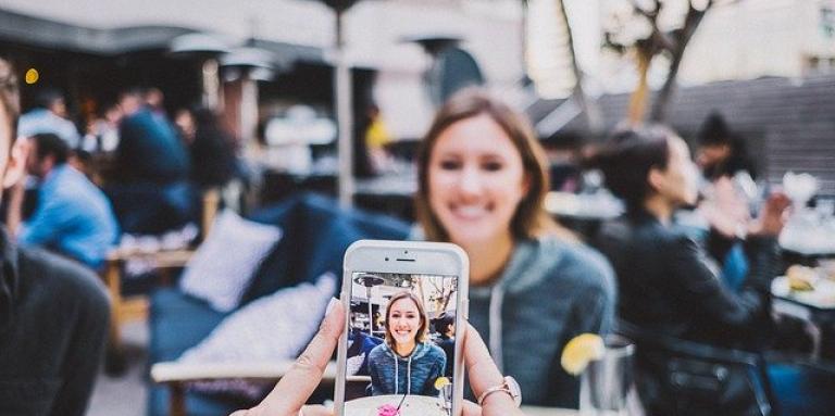 Two women dining. One woman takes a picture of her friend with her smartphone.