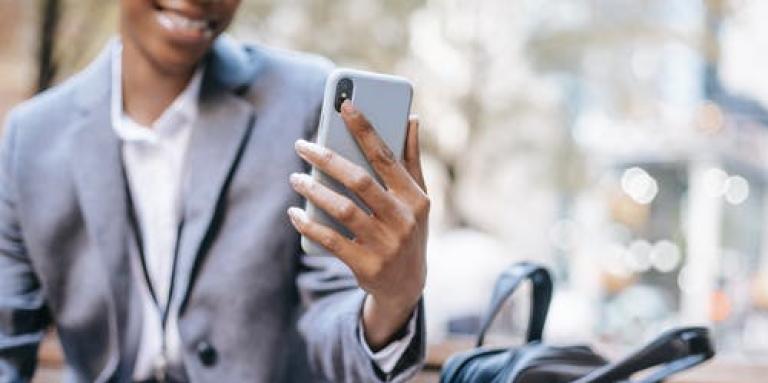 Femme noire assise sur un banc de parc, portant une veste d'affaires et tenant son téléphone intelligent.