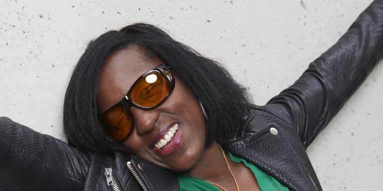 Une femme noire, souriante, portant de grosses lunettes orangées, les bras dans les airs.