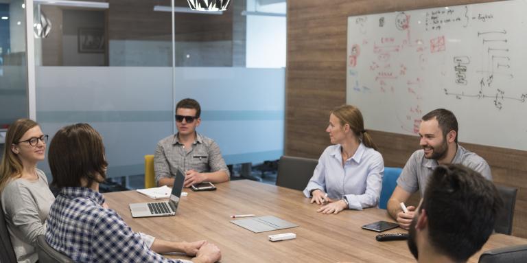 A workplace meeting in a board room. 6 people sit around a long table.