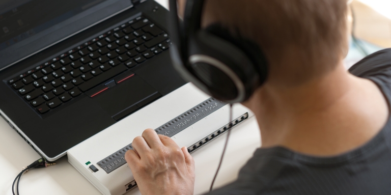 A man is working on laptop computer with a refreshable braille display. He is wearing headphones.