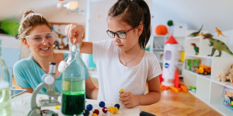 A young girl with glasses stands in front of a table with a microscope and a bottle filled with green liquid.