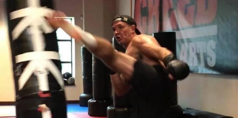 Man wearing black shorts and boxing gloves is in a gym and is doing a high kick to a punching bag.