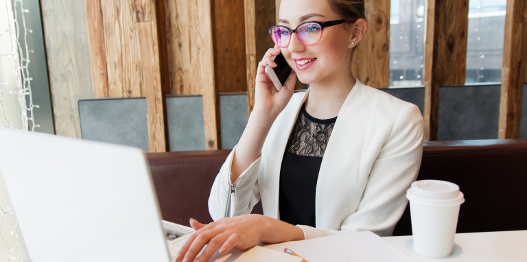 Une jeune femme portant des lunettes parle au téléphone et est sur l'ordi. un café pour emporté est déposé sur la table.